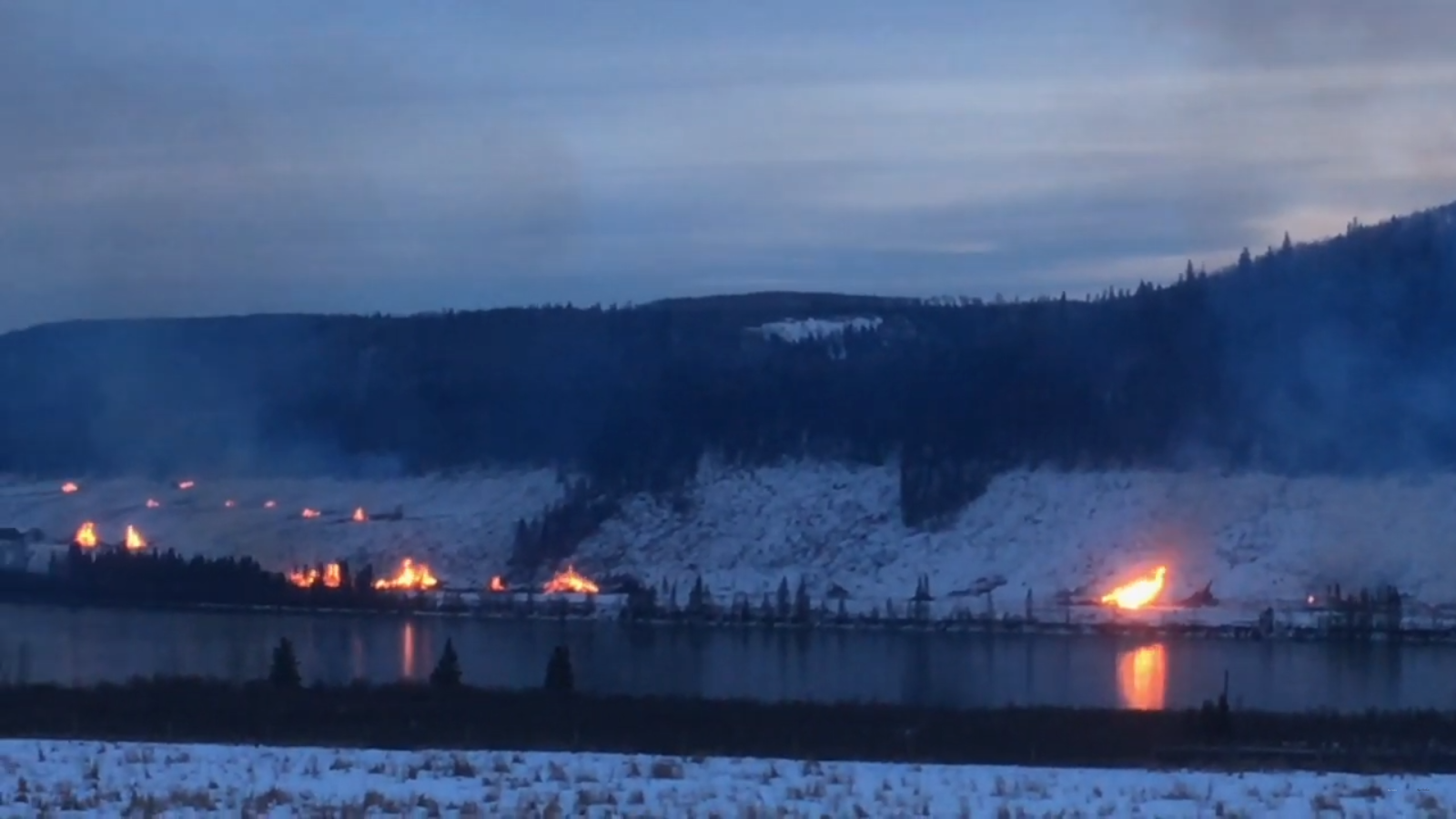 River and hills forested landscape on a dark winter day with snow on the ground, with spot fires burning along the river's edge.